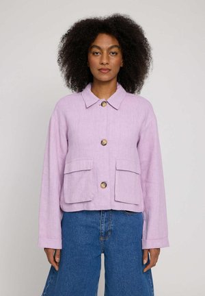 Woman with curly hair wearing a light purple buttoned jacket with two front pockets and blue jeans, standing against a plain background.