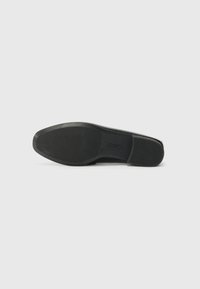 Semelle en caoutchouc noir avec motif de grip texturé, forme légèrement courbée et bord supérieur lisse. Logo de la marque visible sur la semelle.