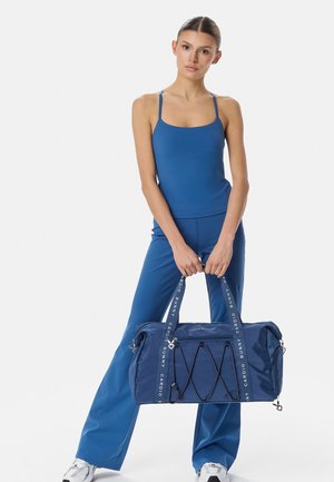 Naine sinises tankitopis ja pükstes, kes hoiab suurt sinist spordikotti, mille kottidel on kaubamärgiga sangad ja mustad elastsed nöörid.