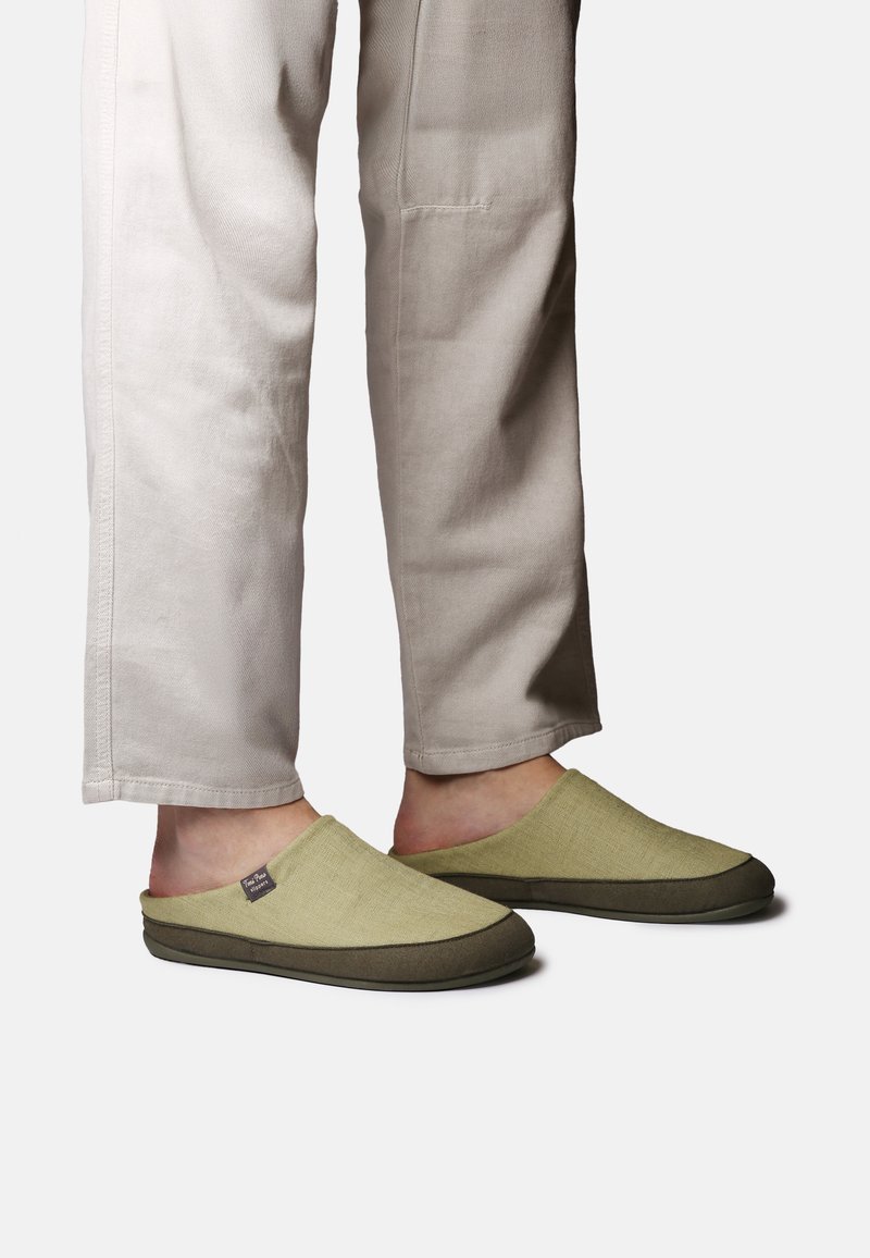 Füße, die beige Schlupfschuhen mit dunklen Sohlen tragen, dazu hellfarbene Hosen, sichtbar von der Mitte der Wade abwärts, vor einem einfarbigen Hintergrund.