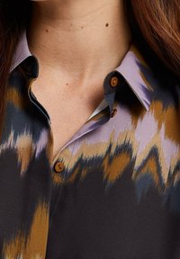 Närbild av en persons nacke och övre bröst som bär en skjorta med krage, med ett flerfärgat abstrakt mönster i lila, brunt, svart och beige.