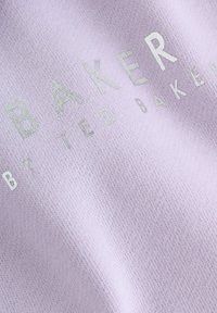 Lila szövet, puha textúrával, fémes ezüst szöveggel, amelyen az áll: "BAKER" és "BY TED BAKER", finom kontrasztot teremtve.