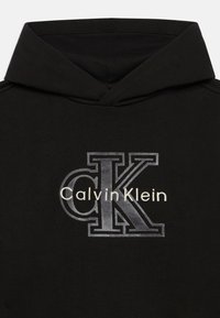 Sweatshirt preto com capuz, feito de um tecido macio, apresentando um design de logótipo em prateado. O logótipo inclui um proeminente "CK" e o texto "Calvin Klein".