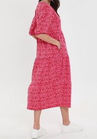 Vestido maxi en rosa con un patrón de zigzag, mangas cortas abullonadas, bolsillos laterales y un corte holgado, combinado con zapatillas blancas.