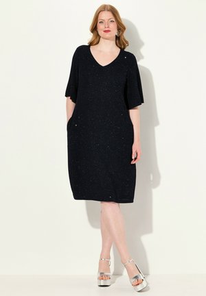 Schwarzes, kurzärmliges Kleid mit V-Ausschnitt und Taschen, das eine gestrickte Textur aufweist. Es reicht bis zur Hälfte der Wade und wird mit metallischen Plateausandalen kombiniert.