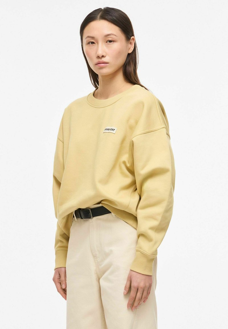 Jeune femme portant un sweat-shirt beige ample avec un petit logo et un pantalon clair avec une ceinture noire, debout devant un fond blanc.