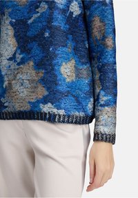 Blauer, floral gemusterter Strickpullover mit beigen und grauen Akzenten, getragen mit hellbeigen Hosen, Nahaufnahme des Oberkörpers und der rechten Hand.