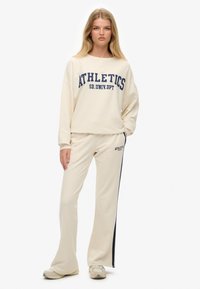Crème-kleurige set van sweatshirt en broek met de tekst "ATHLETICS" in marineblauw. Het sweatshirt heeft verlaagde schouders; de broek heeft een zijstripedetail.