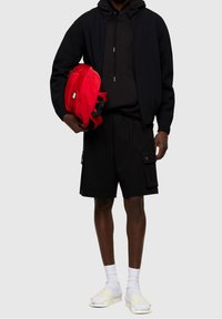 Giubbotto bomber nero sopra un felpa nera con cappuccio, abbinato a pantaloni cargo neri. Zaino rosso, calze bianche e ciabatte color crema.