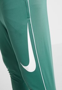 Närbild av gröna sportbyxor med vita sidopaneler och en stor vit swoosh-logotyp på låret.