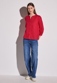 Rotes Hemd mit Knopfleiste und 3/4 Puffärmeln, kombiniert mit hellblauen Straight-Leg-Jeans und weißen Turnschuhen. Glattes Material mit dezentem Glanz.