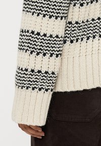 Tloušťkový pletený svetr v krémové barvě s černými vodorovnými pruhy. Má žebrované manžety a lem. Textura látky je silná a útulná.