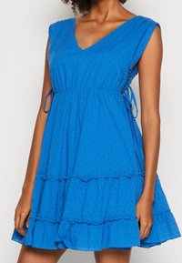 Vestido de algodón azul con escote en V, mangas cortas y detalles de lazo en la cintura. Presenta un dobladillo acampanado con volantes y un patrón texturizado de puntos.