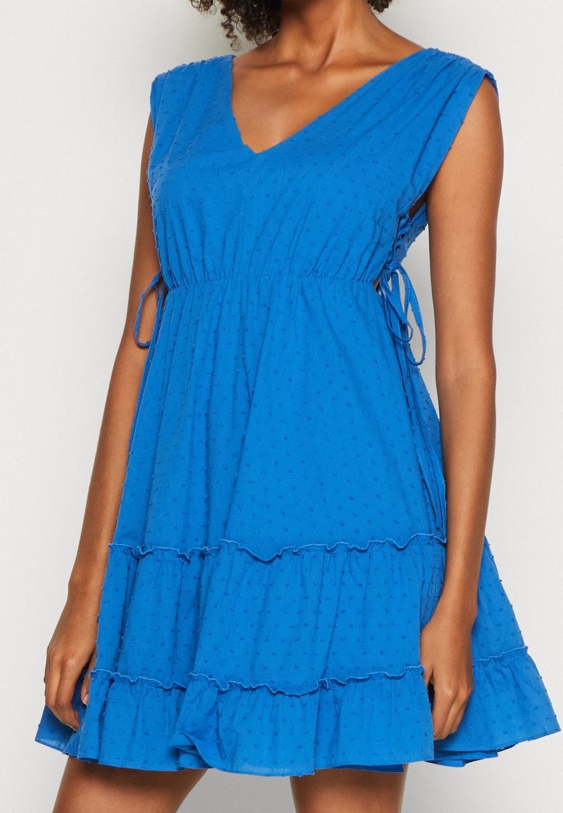 Vestido de algodón azul con escote en V, mangas cortas y detalles de lazo en la cintura. Presenta un dobladillo acampanado con volantes y un patrón texturizado de puntos.