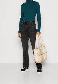 Beige gepolsterte Tragetasche mit zwei Griffen, getragen mit einem grünen Langarmoberteil und dunklen Jeans. Schwarze Sneakers mit weißen Sohlen sind sichtbar.