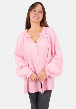 Femme souriante portant un chemisier rose clair ample avec des boutons et une bordure en dentelle sur le col et les poignets, assorti à un pantalon gris foncé, posant devant un fond blanc.