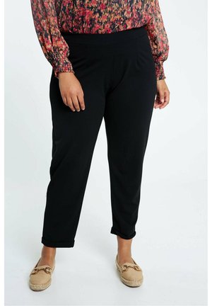 Vrouw die zwarte taps toelopende broek draagt, een bloemenprint blouse met lange mouwen en beige loafers met een gouden gesp, staand op een witte achtergrond.