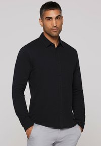 Black long-sleeve shirt made of textured fabric, featuring a button-down collar and front buttons, worn with light gray pants.