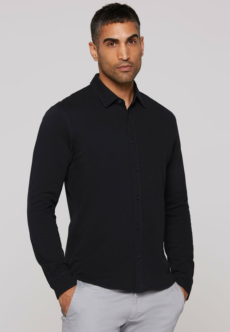 Black long-sleeve shirt made of textured fabric, featuring a button-down collar and front buttons, worn with light gray pants.