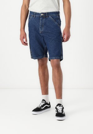 Shorts en denim bleu foncé, présentant une coupe droite, des coutures visibles et des poches latérales, associés à des baskets noires et des chaussettes blanches.
