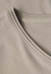 Beige bomulls t-shirt med rund halsringning. Har synliga sömmar och en slät textur, framhävd av naturligt ljus.