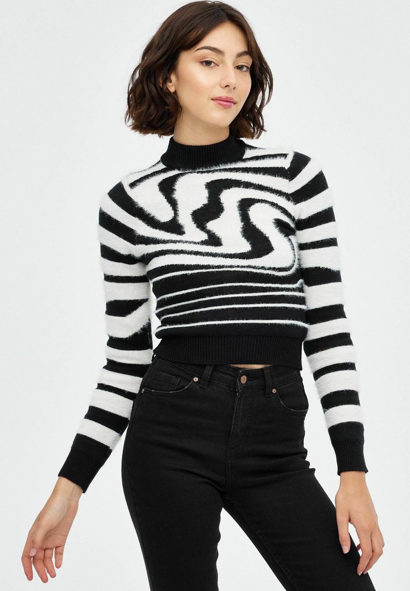 Suéter recortado de rayas en blanco y negro con un cuello alto, textura esponjosa y patrón en espiral. Combinado con pantalones vaqueros negros de cintura alta.