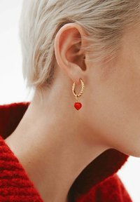 Primer plano de una oreja con cabello rubio corto que lleva un pendiente de aro dorado con un pequeño charm en forma de corazón rojo, frente a un fondo de punto rojo.