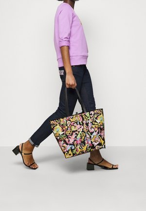 Persoon die loopt en een grote zwarte draagtas met kleurrijke bloem- en krulmotieven draagt, gekleed in een paarse trui, donkere spijkerbroek en zwarte hakken.