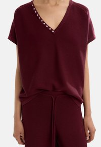 Burgundowa dzianinowa bluzka z krótkimi rękawami i dekoltem w kształcie litery V, ozdobiona białymi guzikami wzdłuż dekoltu. Miękka tekstura, luźny krój.