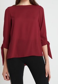 Blusa color bordeaux con scollo quadrato, maniche a tre quarti e dettagli a fiocco ai polsini. Realizzata in tessuto leggero.