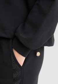 Sweatshirt noir en tissu texturé, avec un logo subtil sur la manche et un cordon de serrage en simili cuir avec un accent doré.