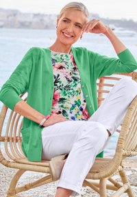 Grüne Strickjacke über einem blumigen, farbenfrohen Top. Weiße Hose und ein hellfarbiges Schuhwerk. Sitzt auf einem geflochtenen Stuhl am Wasser.