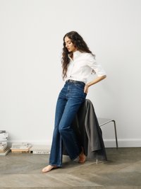 Svetlo modri skinny flare kavbojke z visokim pasom, v kombinaciji z belo srajco s kapuco. Bosi, s sivo jakno, obešeno na stolu.