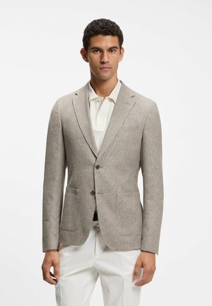 Mann trägt beigen strukturierten Blazer über weißem Poloshirt und weißen Hosen, steht vor schlichtem weißen Hintergrund.