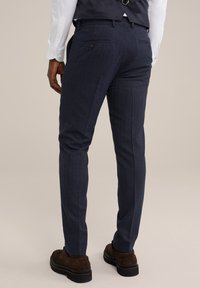Pantalons texturés bleu marine au style ajusté, avec un motif subtil et deux poches arrière avec boutons. Associés à des chaussures marron foncé.