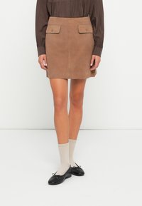 Bruine suede mini-jurk met twee voorkruiszakken, knopenaccenten en een gladde textuur, gecombineerd met zwarte schoenen en beige sokken.