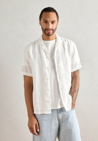 Hvit linskjorte med kort erme og knappelukking, brukt over en hvit t-skjorte, sammen med lyseblå denimjeans.