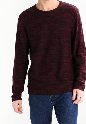 Homme portant un pull à manches longues marbré rouge foncé et noir, associé à un jean bleu, se tenant devant un fond blanc uni.