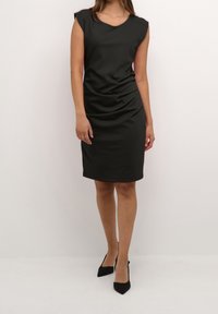 Svart klänning med knälång längd och korta ärmar, texturerad tyg och subtila rynkor på sidan. Matchad med svarta klackskor med spetsig tå.