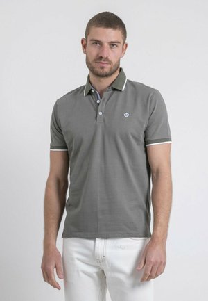 Homme barbu aux cheveux courts portant un polo gris avec bordure blanche et un pantalon blanc, debout devant un fond clair uni.