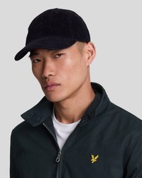 Căciulă din corduroy navy cu o margine curbată și o suprafață texturată, împerecheată cu o jachetă cu fermoar de culoare verde închis și un tricou alb dedesubt.