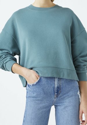 Kvinde iført en løs teal sweatshirt med op-rullede ærmer og lyseblå højtaljede jeans, den ene hånd i lommen mod en ensfarvet baggrund.