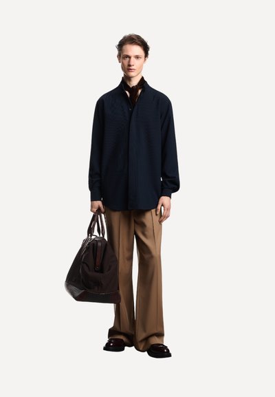Persona con una maglietta nera a maniche lunghe, pantaloni beige a gamba larga, mocassini scuri, che tiene una grande borsa da viaggio in pelle e tessuto marrone.