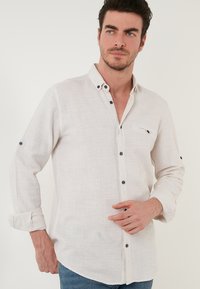 Világos bézs, hosszú ujjú gombos ing textúrált anyagból, zsebbel, sötét gombokkal és felgöngyölt ujjakkal. Hétköznapi kialakítás.