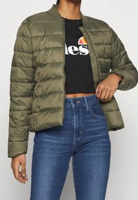 Chaqueta acolchada en color verde oliva, con cierre de cremallera y cuello acanalado, combinada con una camiseta negra corta y jeans azules de cintura alta.