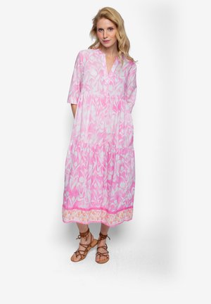 Różowa sukienka w kwiatowy wzór z warstwowym fasonem, krótkimi rękawami i dekoltem w serek, z kontrastowym ozdobnym brzegiem. Stylizacja dopełniona sandałami.