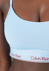 Lichtblauwe bralette gemaakt van rekbare stof, met een brede witte band waarop "Calvin Klein" in rode letters staat. Ontworpen met dunne bandjes.
