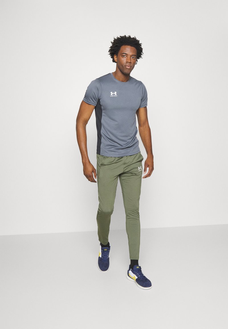 T-shirt de sport gris à manches courtes, associé à un pantalon de jogging vert. Chaussures de couleur bleu foncé avec des accents jaunes. Design simple et fonctionnel.