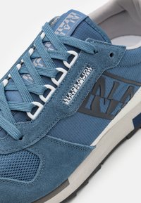 Sneaker blu con tomaia in suede e tessuto, caratterizzato da rete testurizzata, design a strati, lacci bianchi e un logo prominente sul lato.