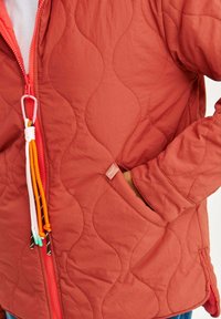 Czerwona pikowana kurtka z zamkiem błyskawicznym z przodu, z falistym wzorem, regulowanymi akcentami sznurków oraz bocznymi kieszeniami. Teksturowana tkanina o gładkim wykończeniu.